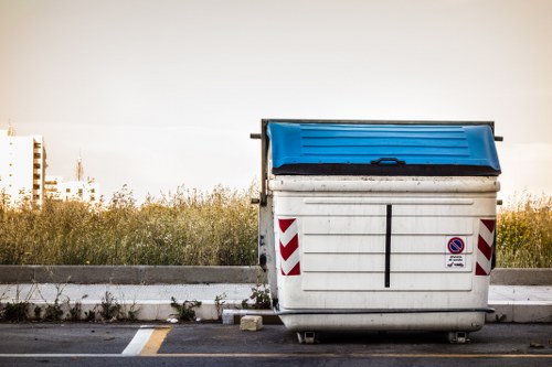 A large commercial rubbish skip with a white body, vertical metal slats, and red and white striped reflective markings positioned on a paved surface. The skip has a blue, hinged lid on top, slightly tilted forward. It features a small sign on the side indicating no parking, with a blue circle and red slash. The background shows a low curb, some overgrown grass and wild plants, and a clear sky with a faint horizon line, suggesting an outdoor setting near a South Suffolk area, possibly Sudbury. The overall scene is well-lit with natural daylight, emphasizing the utilitarian appearance typical of waste collection containers used in commercial or office clearance services.