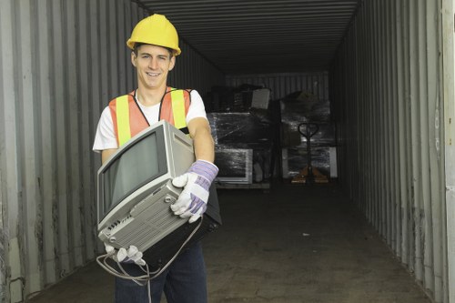 A young man wearing a yellow safety helmet, white t-shirt, high-visibility vest with reflective orange and yellow strips, and work gloves is standing in the entrance of a large grey metal storage container or shipping container. He is smiling and holding an old, beige CRT computer monitor with a visible power cord hanging down. Inside the container, there are stacked black plastic-wrapped electronic waste or debris, which appear to be discarded appliances or equipment. The container is situated on a concrete or paved surface, likely part of a commercial or industrial area in Sudbury or nearby. The lighting is natural, suggesting daytime, and the overall scene relates to waste removal or clearance services, reflecting the type of rubbish commonly handled by Office Clearance Sudbury.