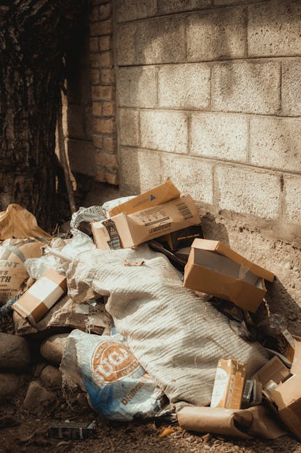 A pile of various waste materials including flattened and partially opened cardboard boxes, plastic bags, and paper packaging is situated outdoors against a rough concrete wall with visible blocks and