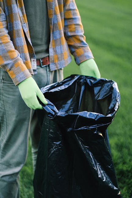 A pile of various waste materials including flattened and partially opened cardboard boxes, plastic bags, and paper packaging is situated outdoors against a rough concrete wall with visible blocks and