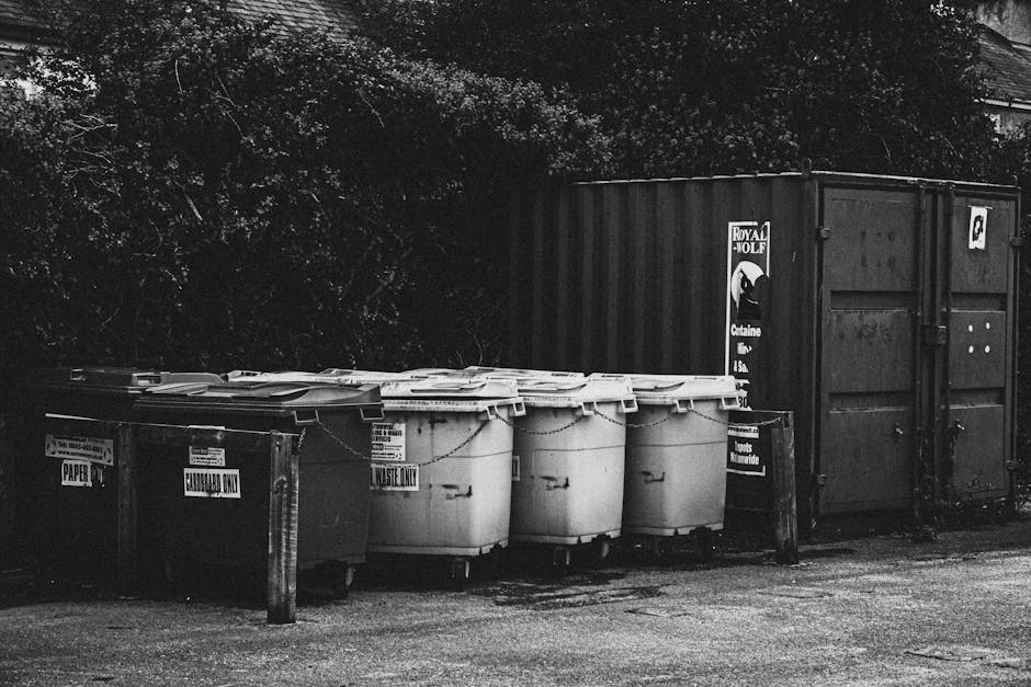 A row of four large, white plastic wheelie bins with closed lids, positioned on a paved surface in front of a dark, industrial-style shipping container. The bins each have labels indicating they are f