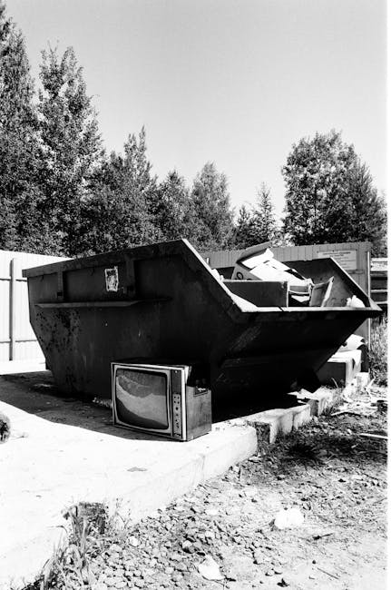 A large pile of assorted waste materials is stacked on a paved driveway outside a residential building. The collection includes wooden planks with rough, weathered surfaces in shades of brown and beig