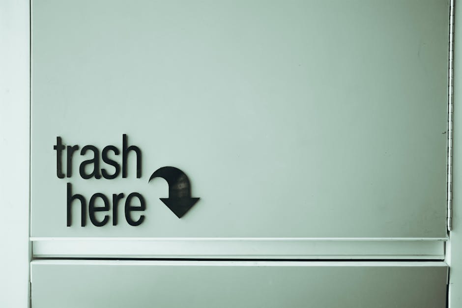 A close-up view of a light-colored, smooth wall with a black sign that reads 'trash here' in lowercase letters, accompanied by a downward-pointing arrow, indicating a designated rubbish disposal area.